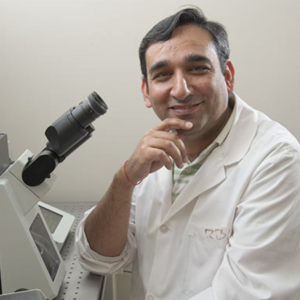 Photo of Dr. Pavneesh Madan wearing a white lab coat and sitting infront of a microscope